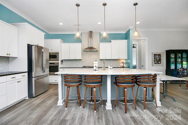 kitchen with hanging light fixtures, wall chimney exhaust hood, stainless steel appliances, white cabinets, and a kitchen island with sink