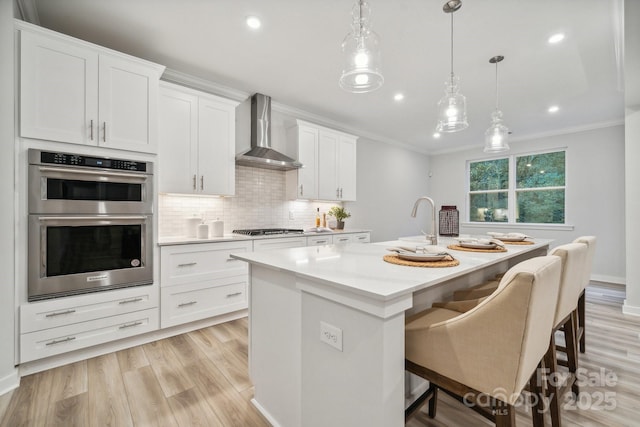 kitchen with hanging light fixtures, a kitchen island with sink, wall chimney range hood, white cabinets, and stainless steel appliances