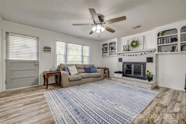 living room with a brick fireplace, a textured ceiling, ceiling fan, crown molding, and light hardwood / wood-style floors