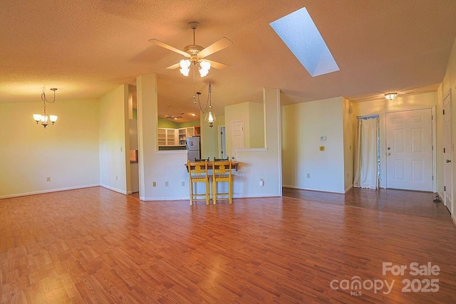 unfurnished living room with a textured ceiling, vaulted ceiling with skylight, ceiling fan with notable chandelier, wood finished floors, and baseboards