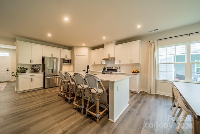 kitchen with appliances with stainless steel finishes, tasteful backsplash, a kitchen island with sink, light hardwood / wood-style flooring, and white cabinets