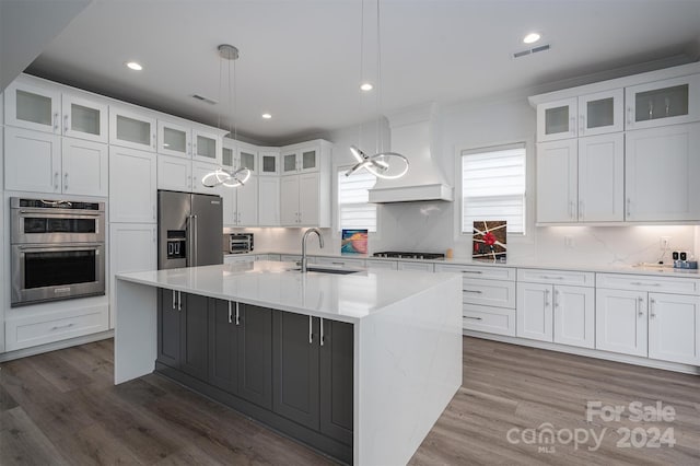 kitchen with pendant lighting, sink, custom exhaust hood, stainless steel appliances, and a center island with sink