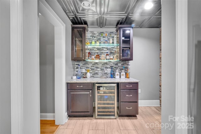 bar featuring wine cooler, backsplash, dark brown cabinetry, and light stone countertops