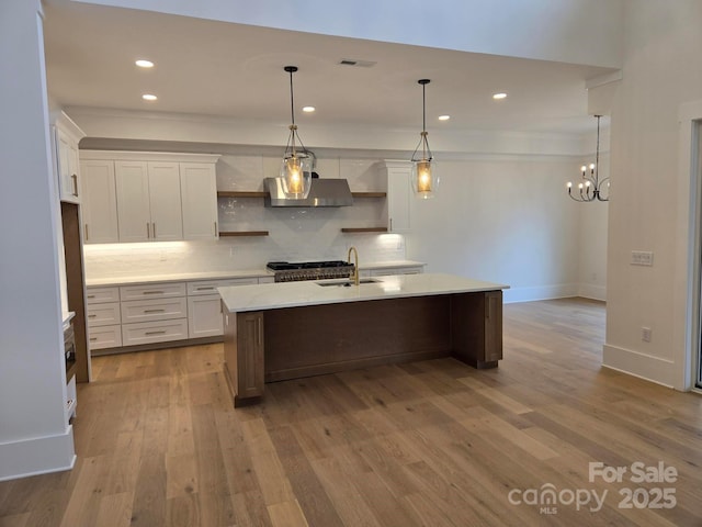 kitchen with decorative light fixtures, sink, white cabinets, a kitchen island with sink, and wall chimney exhaust hood