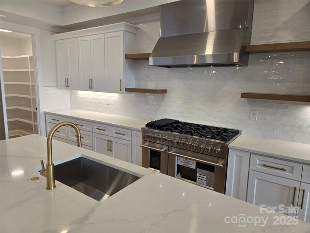 kitchen featuring light stone counters, white cabinets, range with two ovens, and wall chimney range hood