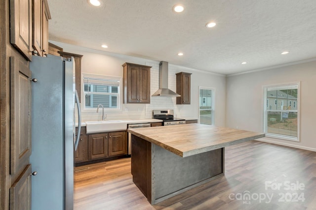 kitchen with appliances with stainless steel finishes, a kitchen island, wall chimney range hood, butcher block counters, and sink