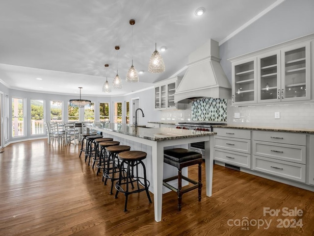 kitchen featuring a center island with sink, backsplash, lofted ceiling, hanging light fixtures, and custom range hood