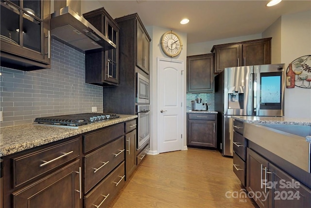 kitchen featuring stainless steel appliances, wall chimney range hood, tasteful backsplash, light stone counters, and light wood-type flooring