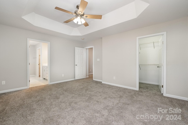 unfurnished bedroom featuring a raised ceiling, a spacious closet, ensuite bath, light colored carpet, and ceiling fan