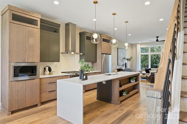 kitchen with ceiling fan, wall chimney range hood, decorative light fixtures, a center island with sink, and appliances with stainless steel finishes