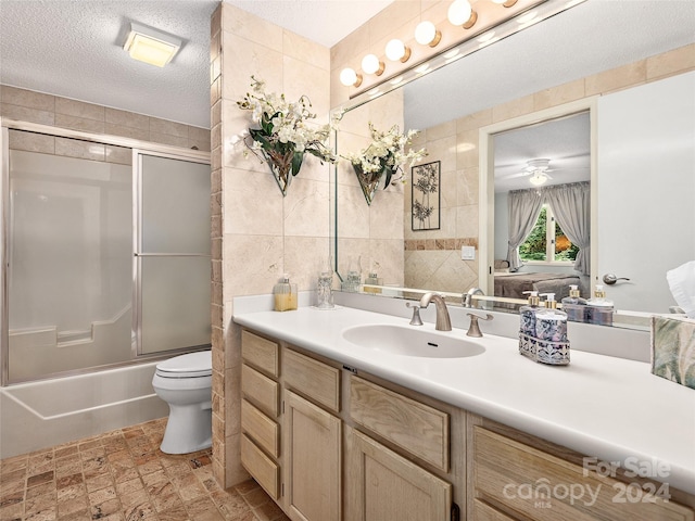 full bathroom featuring vanity, bath / shower combo with glass door, toilet, a textured ceiling, and tile walls