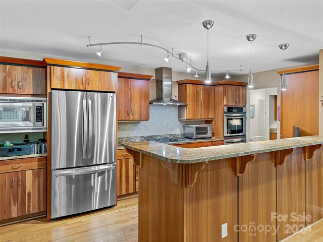 kitchen with appliances with stainless steel finishes, decorative light fixtures, rail lighting, light wood-type flooring, and wall chimney exhaust hood