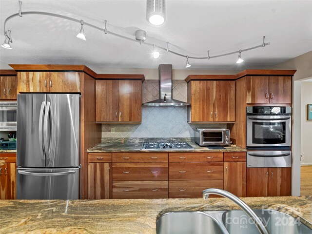 kitchen featuring stainless steel appliances, stone counters, decorative backsplash, wall chimney exhaust hood, and track lighting