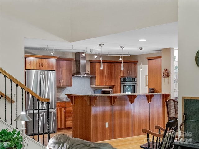 kitchen with light wood-type flooring, kitchen peninsula, stainless steel appliances, wall chimney exhaust hood, and decorative backsplash