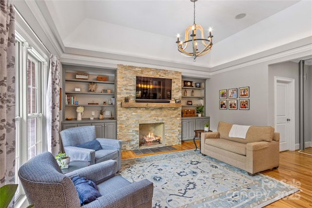 living room featuring a stone fireplace, an inviting chandelier, a tray ceiling, wood-type flooring, and built in shelves