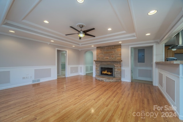 unfurnished living room with ceiling fan, a raised ceiling, a stone fireplace, light hardwood / wood-style flooring, and crown molding