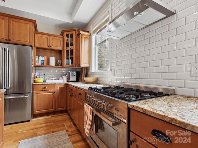 kitchen with decorative backsplash, light stone counters, wall chimney exhaust hood, stainless steel appliances, and light hardwood / wood-style flooring