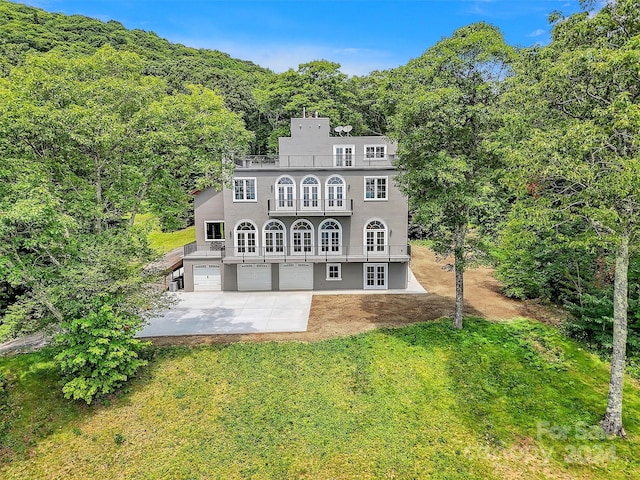 rear view of house featuring a balcony, a forest view, concrete driveway, and a yard