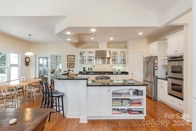 kitchen featuring appliances with stainless steel finishes, a kitchen island with sink, wall chimney range hood, decorative light fixtures, and white cabinets