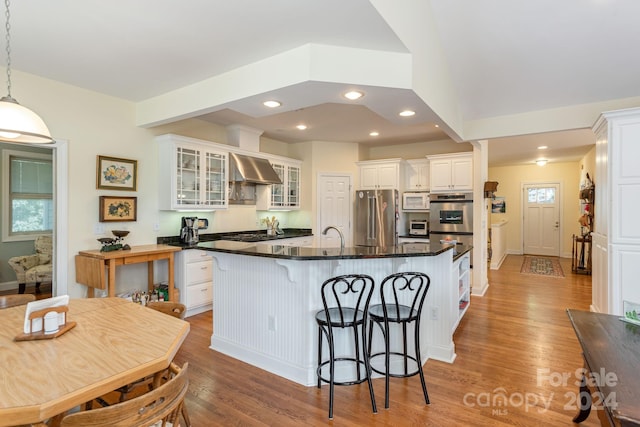 kitchen with white cabinets, a center island with sink, hanging light fixtures, wall chimney exhaust hood, and appliances with stainless steel finishes