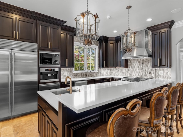 kitchen with wall chimney exhaust hood, decorative light fixtures, built in appliances, sink, and a breakfast bar area