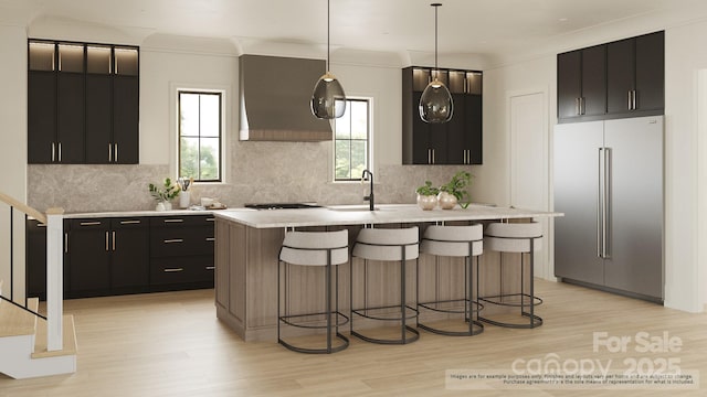 kitchen with stainless steel built in refrigerator, a kitchen island with sink, custom range hood, pendant lighting, and light hardwood / wood-style floors