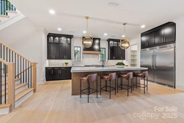 kitchen featuring stainless steel built in fridge, dark cabinets, custom range hood, and light wood finished floors
