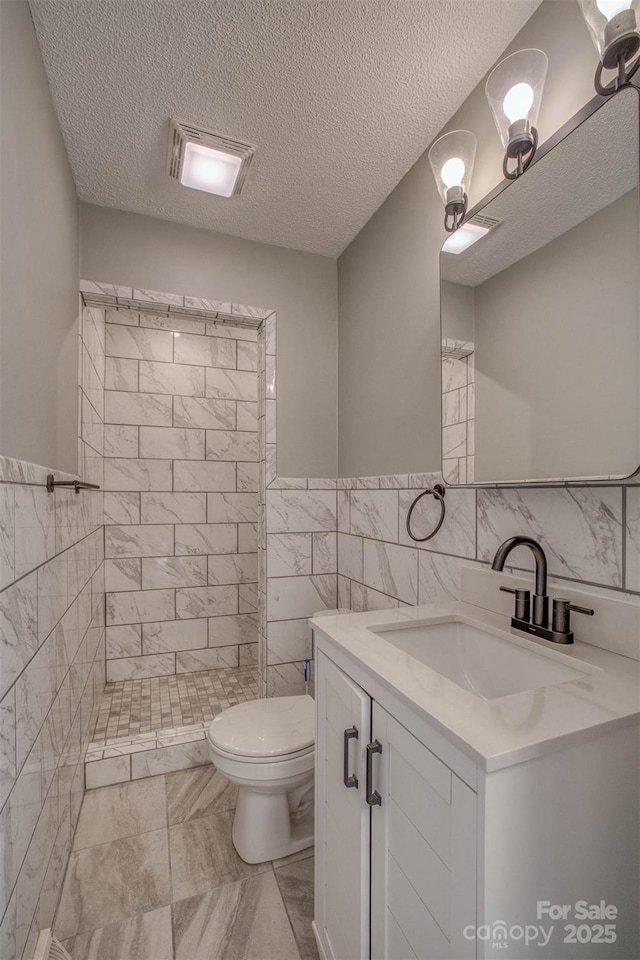 bathroom with a tile shower, tile walls, vanity, toilet, and a textured ceiling