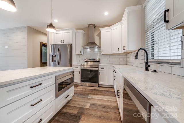 kitchen with wall chimney range hood, white cabinetry, stainless steel appliances, light stone counters, and decorative light fixtures