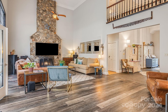 living room with ceiling fan, hardwood / wood-style floors, a high ceiling, a stone fireplace, and ornamental molding