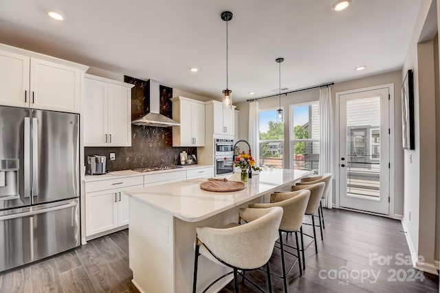 kitchen featuring a center island with sink, appliances with stainless steel finishes, wall chimney range hood, pendant lighting, and white cabinets