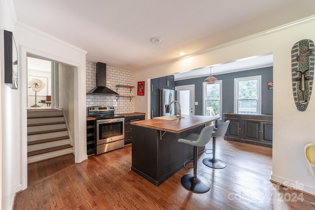 kitchen featuring wall chimney exhaust hood, stainless steel appliances, a center island with sink, butcher block countertops, and dark hardwood / wood-style flooring