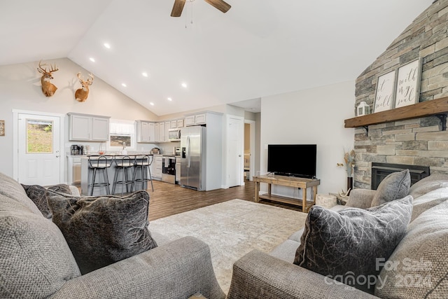 living room featuring a ceiling fan, high vaulted ceiling, a stone fireplace, and light wood finished floors