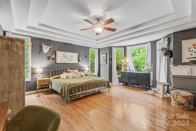 bedroom with ceiling fan, a textured ceiling, a tray ceiling, and light hardwood / wood-style flooring