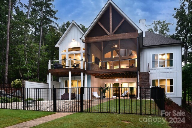 back of property at dusk featuring a lawn, a balcony, a chimney, fence, and board and batten siding