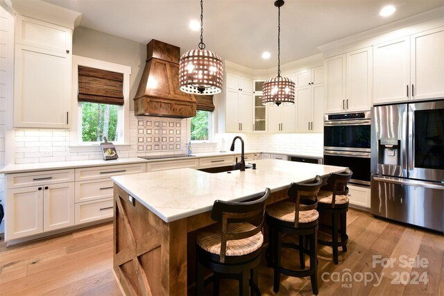 kitchen with a center island with sink, custom range hood, glass insert cabinets, stainless steel appliances, and white cabinetry