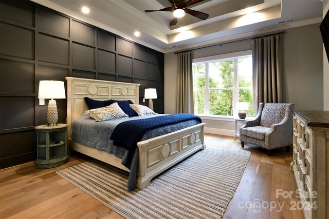 bedroom with ceiling fan, light wood-style flooring, a decorative wall, visible vents, and a raised ceiling