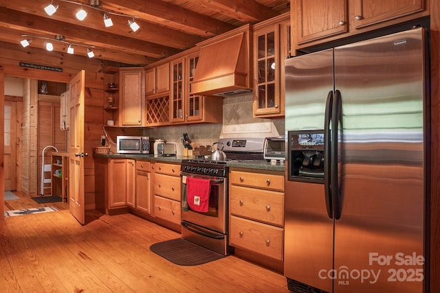 kitchen featuring beam ceiling, dark countertops, premium range hood, light wood-style flooring, and appliances with stainless steel finishes