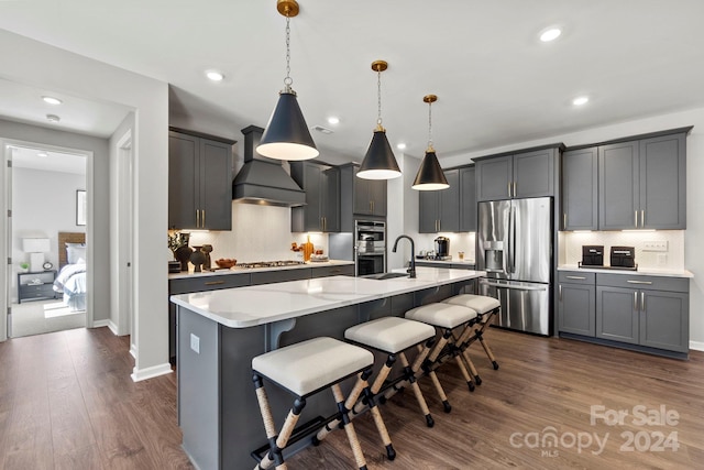 kitchen with custom exhaust hood, stainless steel appliances, tasteful backsplash, a kitchen island with sink, and hanging light fixtures