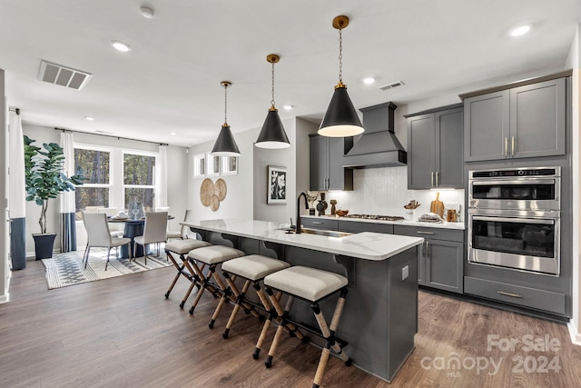 kitchen with stainless steel appliances, an island with sink, sink, hanging light fixtures, and custom range hood