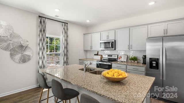 kitchen with an island with sink, stainless steel appliances, decorative backsplash, light stone countertops, and sink