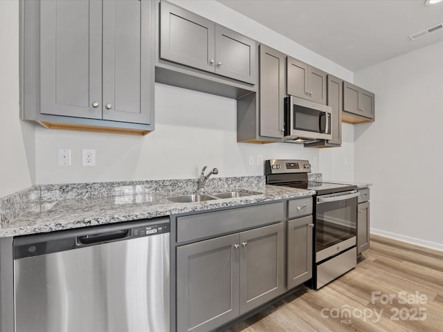 kitchen with gray cabinets, stainless steel appliances, light wood-type flooring, light stone countertops, and sink