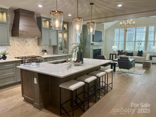 kitchen with a center island with sink, sink, custom exhaust hood, light stone counters, and hanging light fixtures