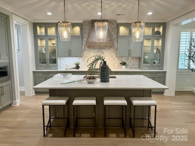 kitchen featuring a center island with sink, pendant lighting, wall chimney exhaust hood, and light stone counters