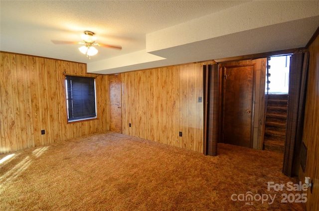 carpeted empty room with stairs, wooden walls, a ceiling fan, and a textured ceiling