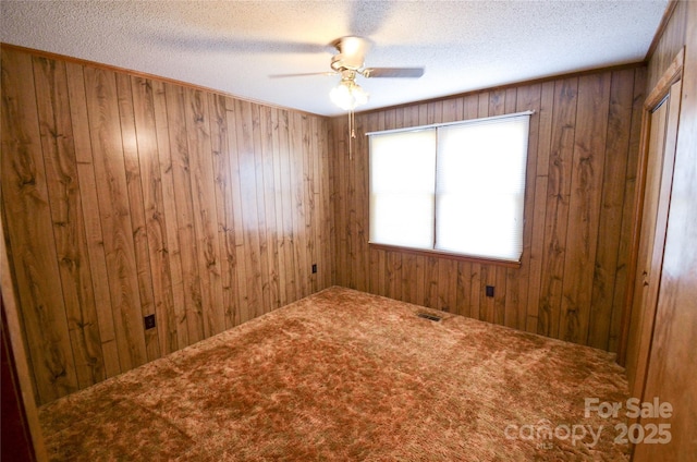 carpeted empty room featuring a textured ceiling, visible vents, a ceiling fan, and wooden walls