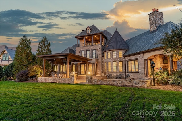 back house at dusk featuring a balcony, a patio area, and a lawn