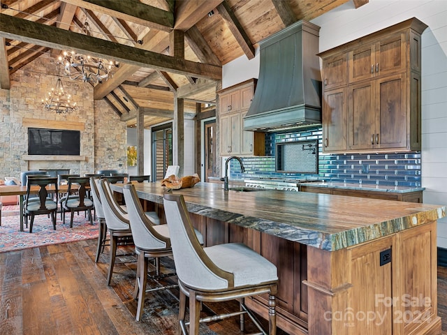 kitchen with dark wood-type flooring, custom exhaust hood, beam ceiling, a kitchen island with sink, and backsplash