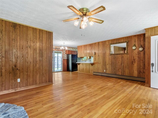 unfurnished living room with light wood-type flooring, baseboard heating, ceiling fan with notable chandelier, a textured ceiling, and wooden walls