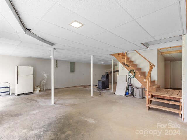 basement featuring a paneled ceiling, white refrigerator, and electric panel